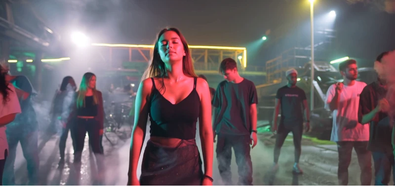 Een vrouw in een zwarte outfit staat zelfverzekerd met gesloten ogen in het midden van een donker verlichte dansscène met neonkleuren, omringd door mensen en mist.